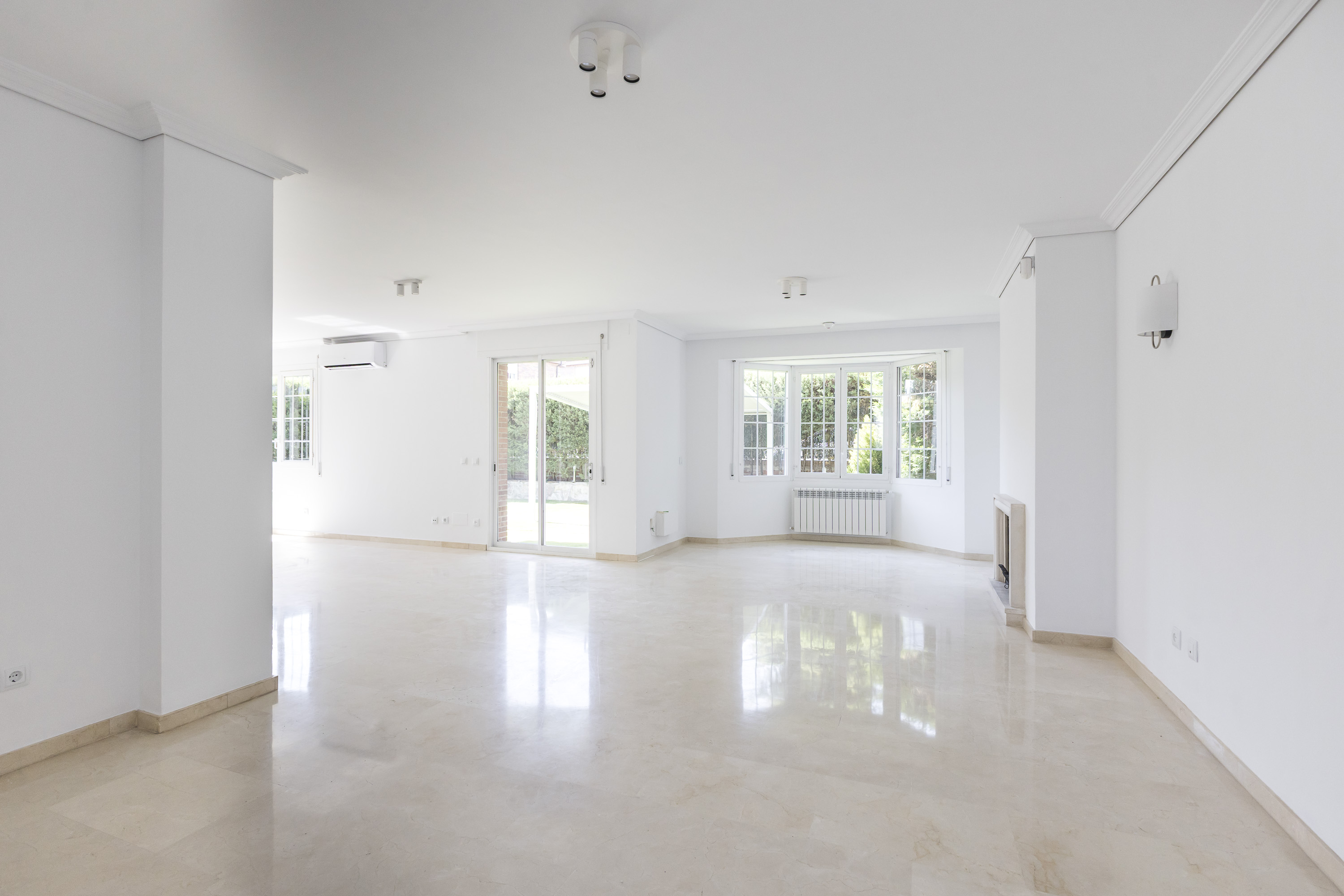 Spacious, bright, empty living room with white walls, polished marble floors, large windows, and a glass door to a garden.