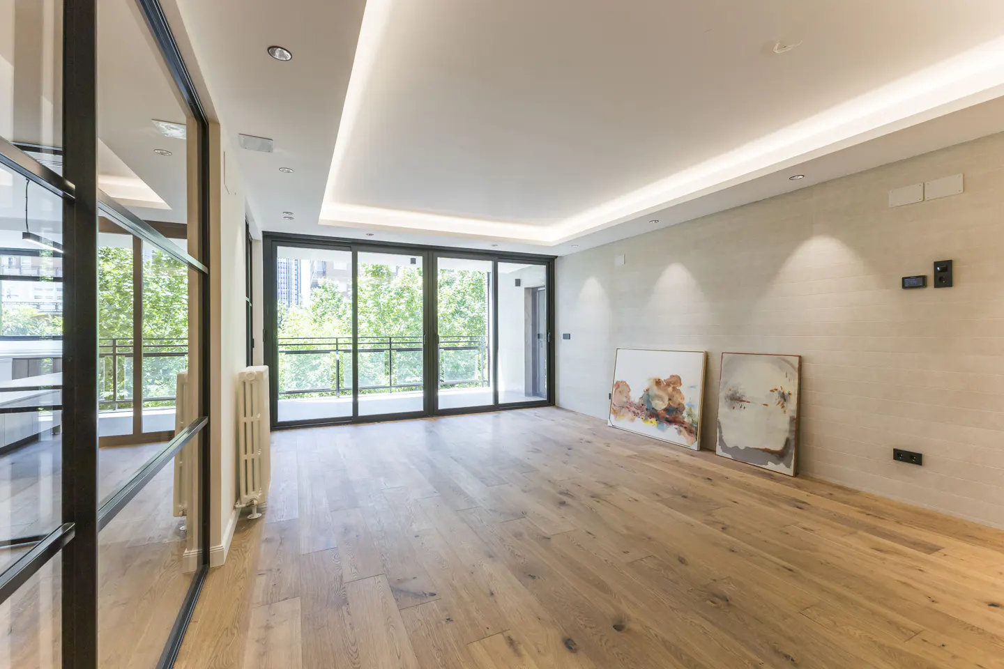 Bright, empty room with wood floors, light walls, and black-framed glass doors to a balcony with trees. Two paintings lean against the wall.