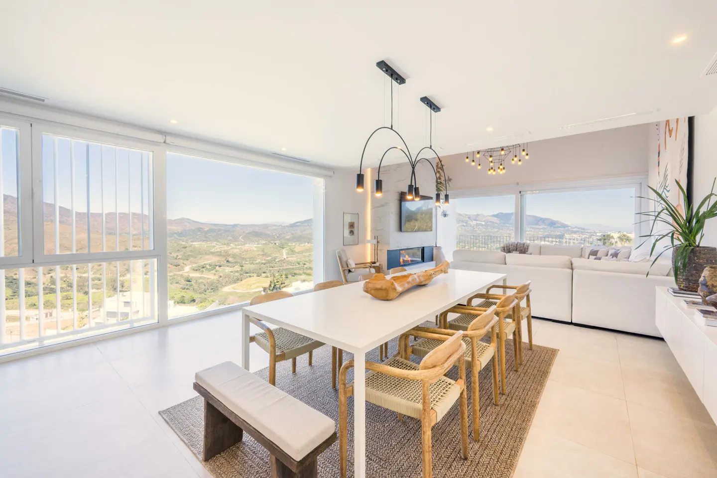 Bright, modern dining room with white table, wood chairs, and bench on a jute rug. Large windows offer a mountain view.