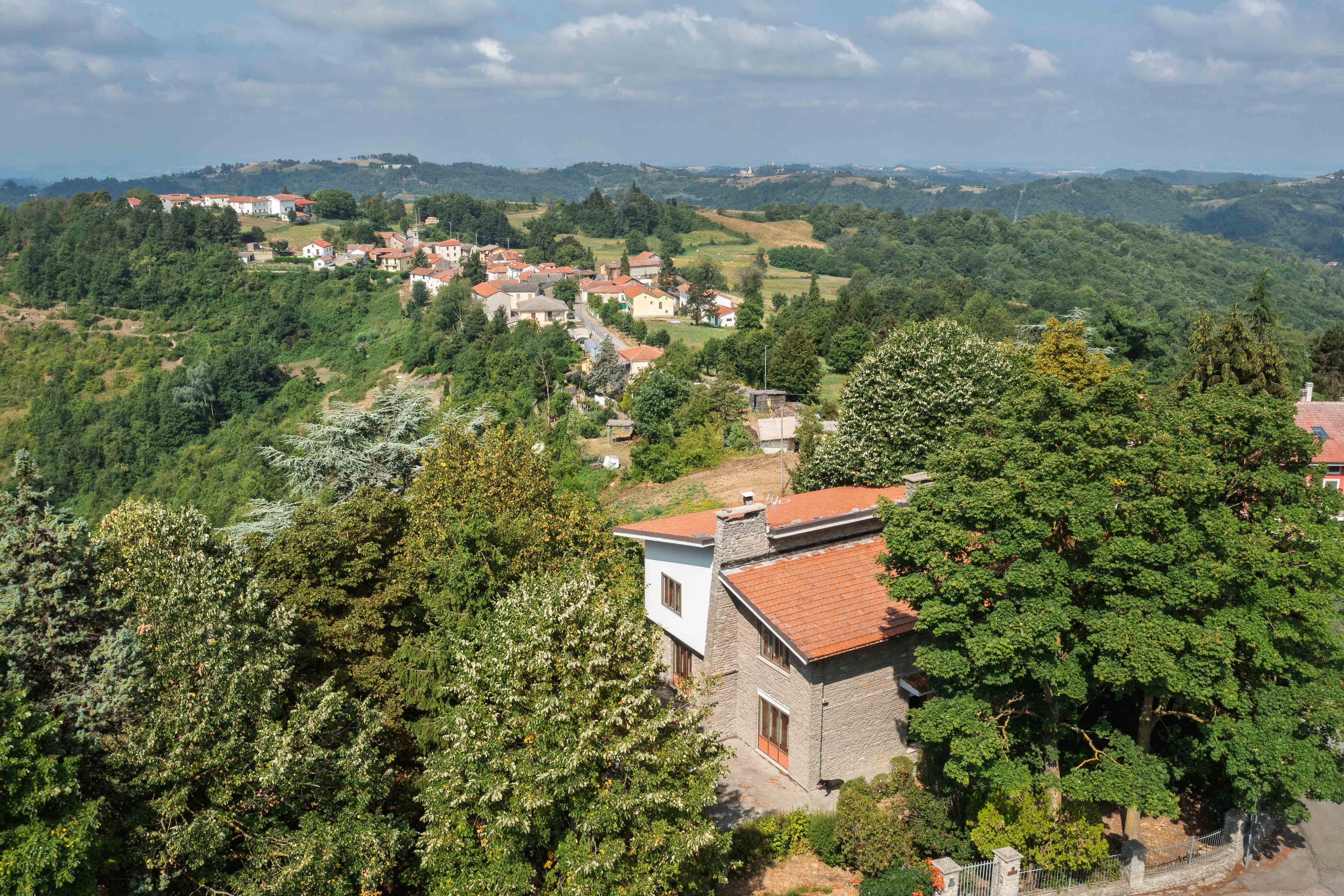 Aerial view of a stone house with a red roof nestled among lush trees, overlooking a scenic hillside village under blue sky. [AI-generated text]
