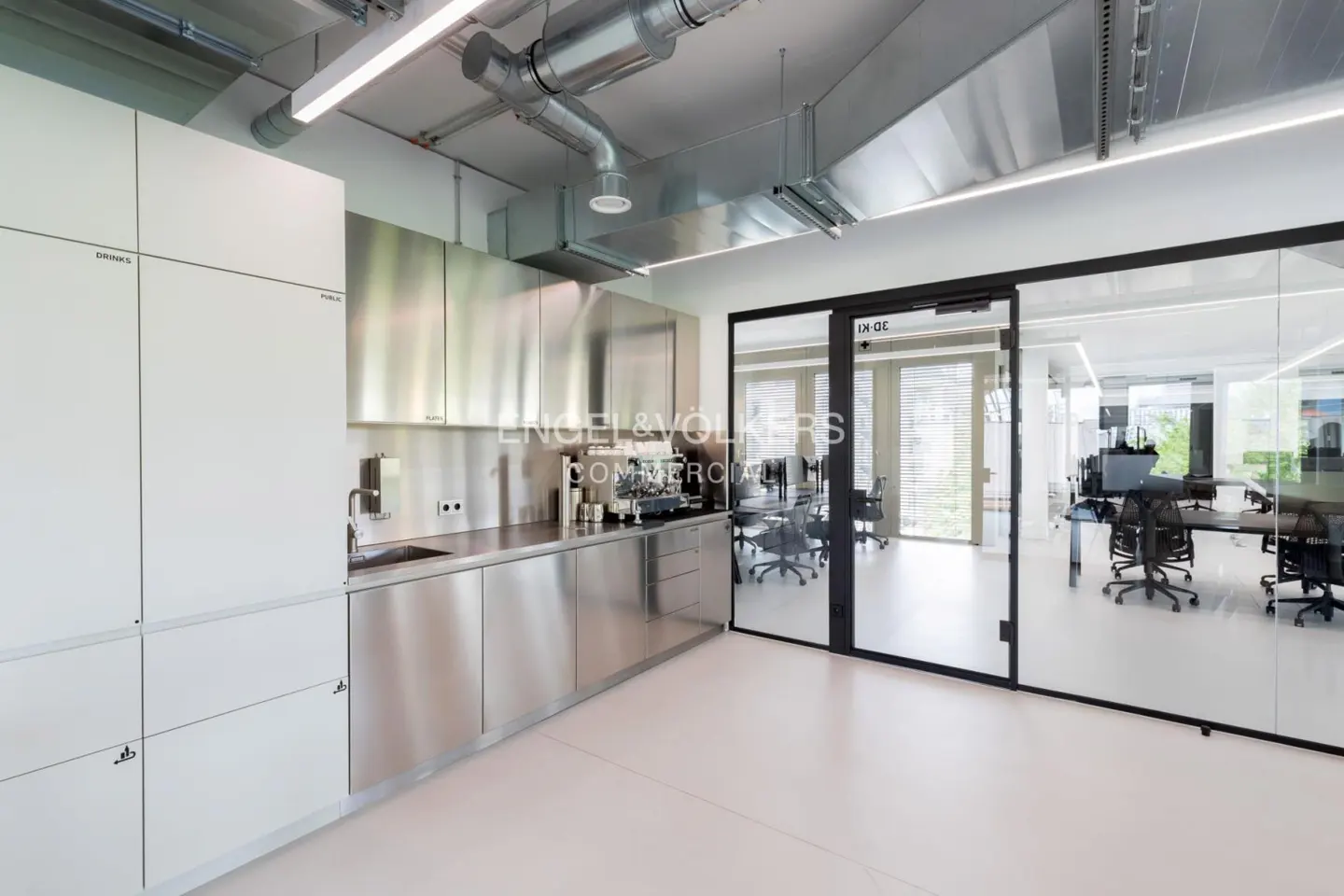 Modern office space with stainless steel kitchen area and glass-walled conference room. White cabinets and light floors create a clean, bright aesthetic.