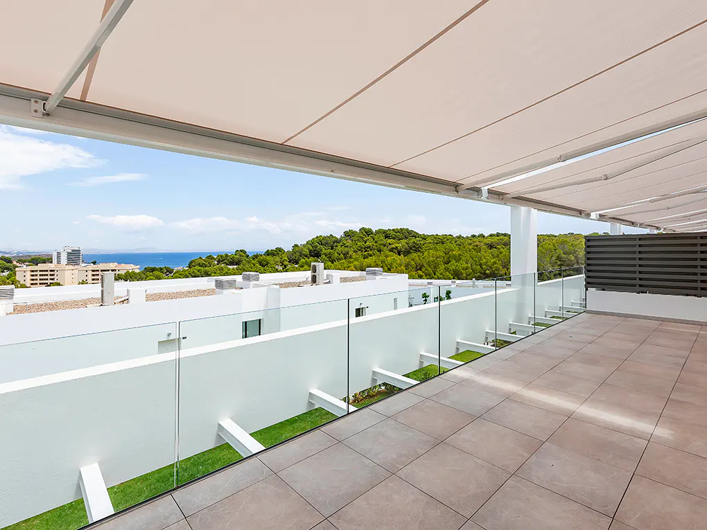 A modern balcony with beige tiles, a glass railing, and a retractable awning. In the background, there's a view of the sea and green trees.