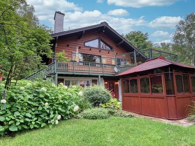 Two-story log cabin with a stone chimney, a balcony, and a red gazebo on a green lawn.