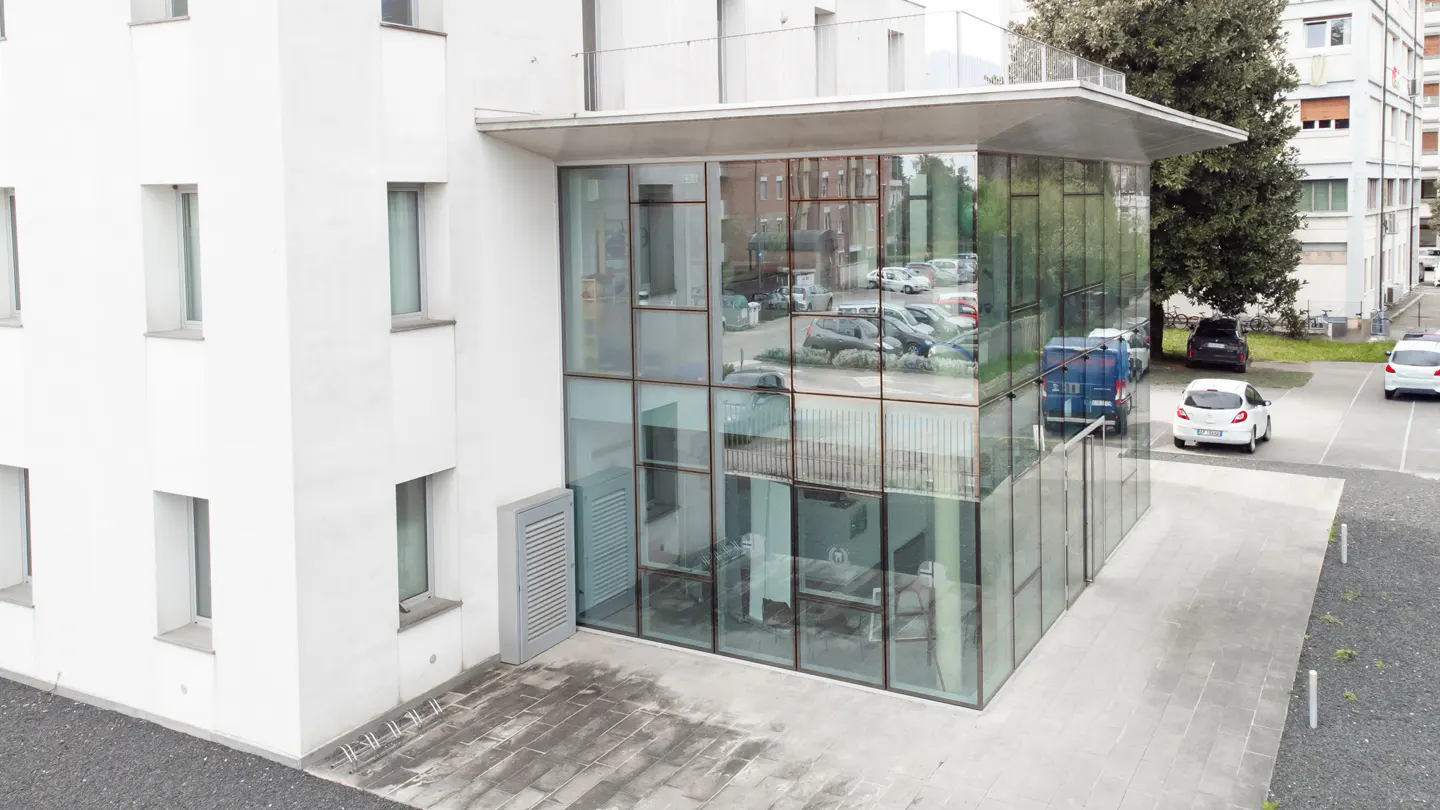 Modern white building with a glass-walled corner extension, reflecting a parking lot with cars on a paved surface.