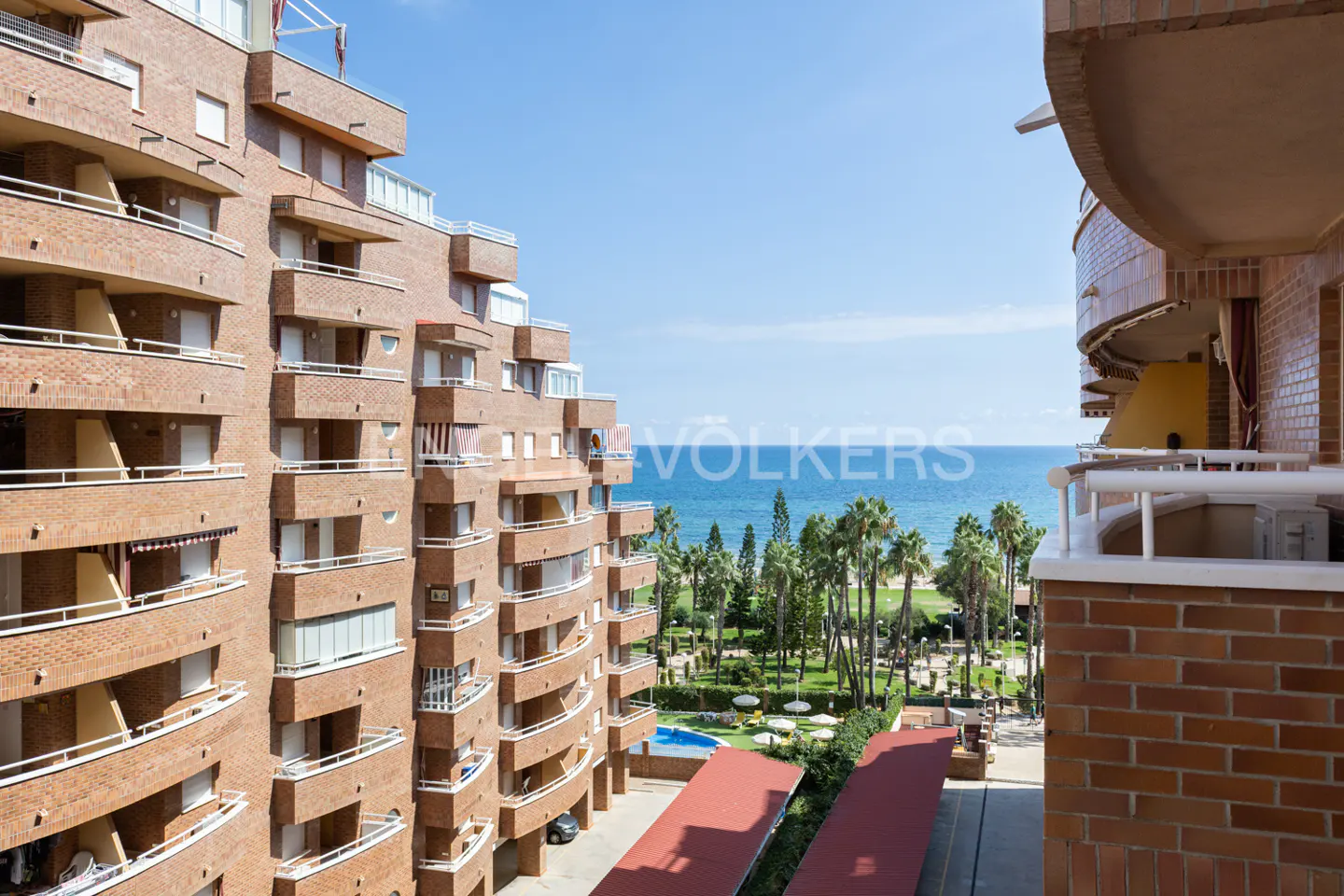 View from a balcony of a brick apartment building overlooking a park, the ocean, and a blue sky.