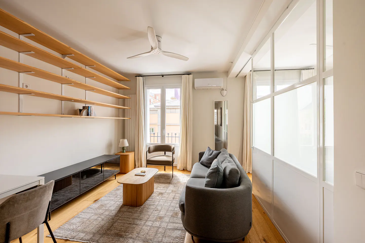 Bright living room with wood floors, gray sofa, and light wood shelving. A window with white curtains is visible.