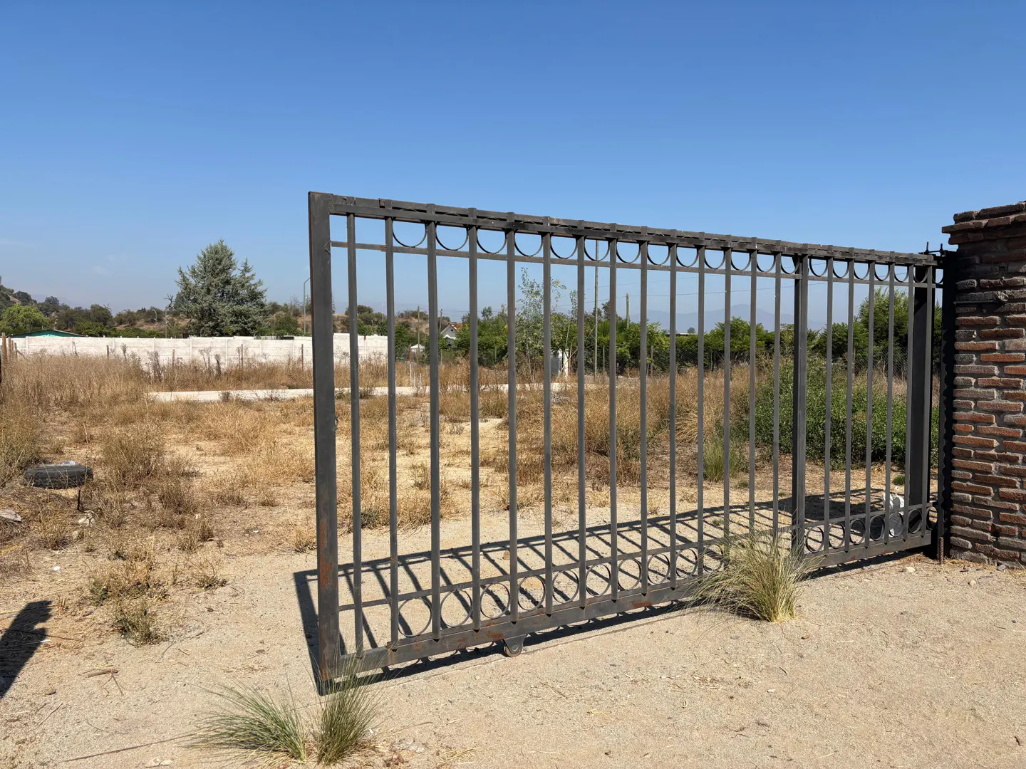 Open wrought iron gate to a vacant lot with dry grass under a clear blue sky.
