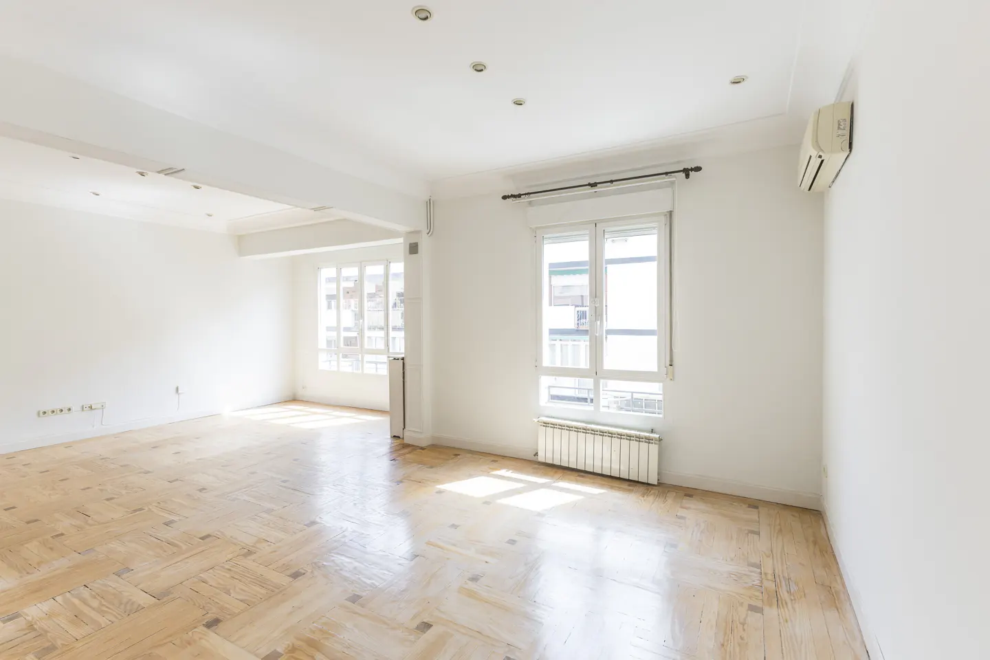 Bright, empty room with parquet floors, white walls, and two large windows letting in sunlight. A white air conditioner is mounted high on the wall.