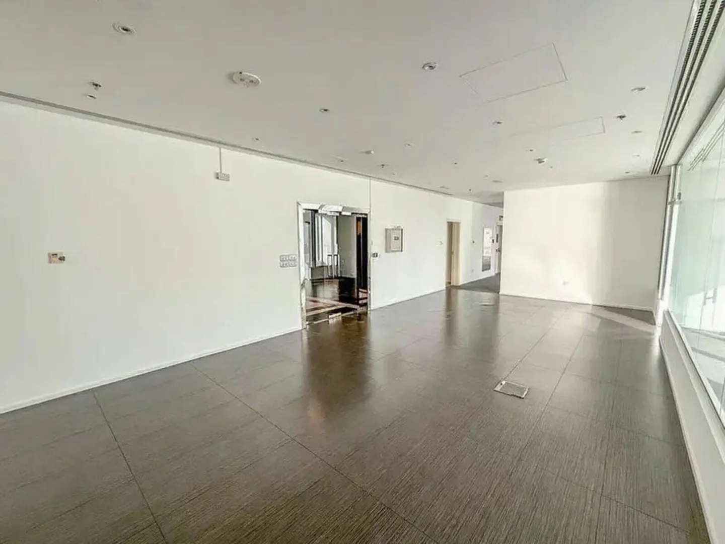 Bright, empty office space with white walls, dark wood-look floors, and large windows on the right.