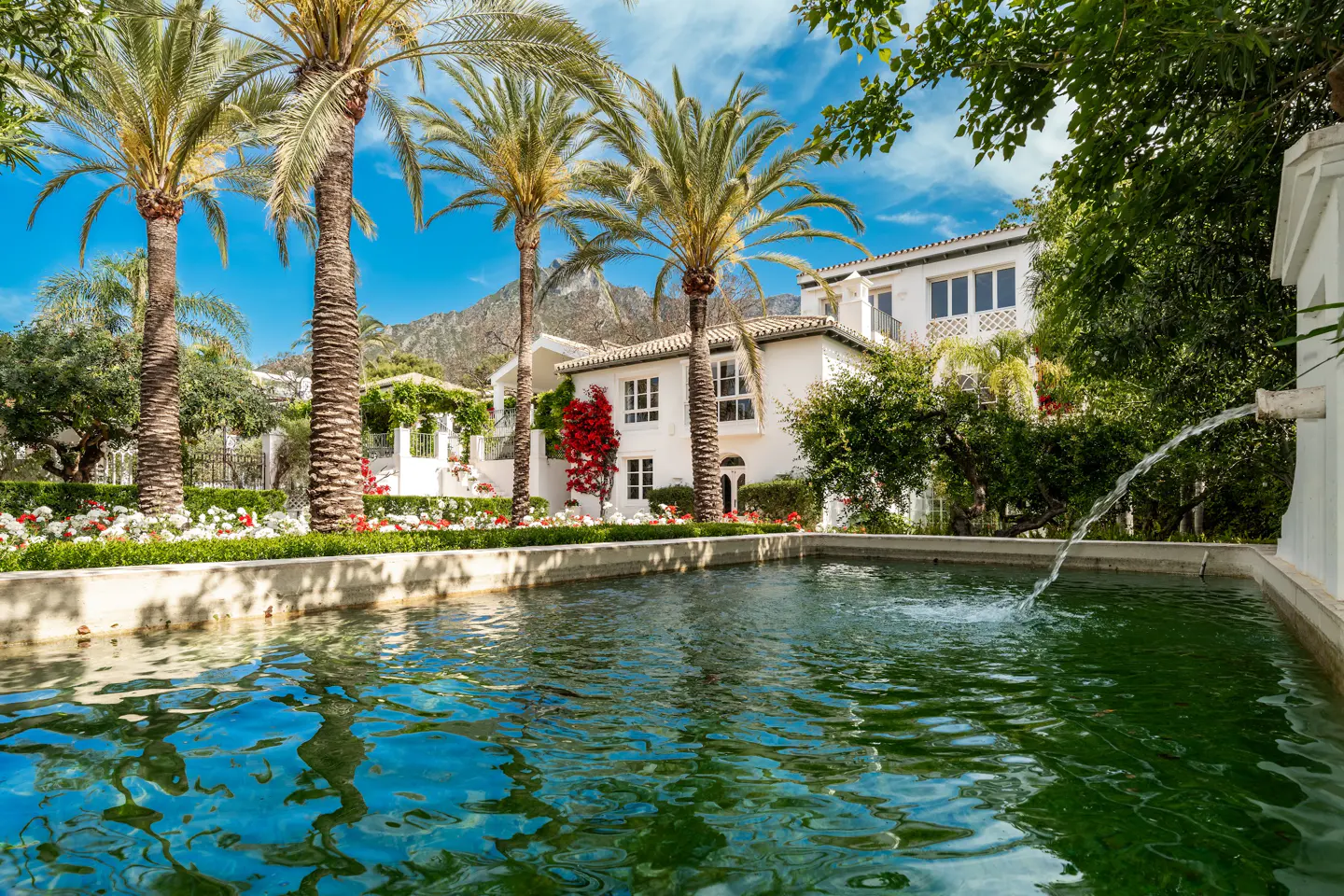 Luxury home with a pool and palm trees. Water flows from a fountain into the pool. Mountains are in the background.