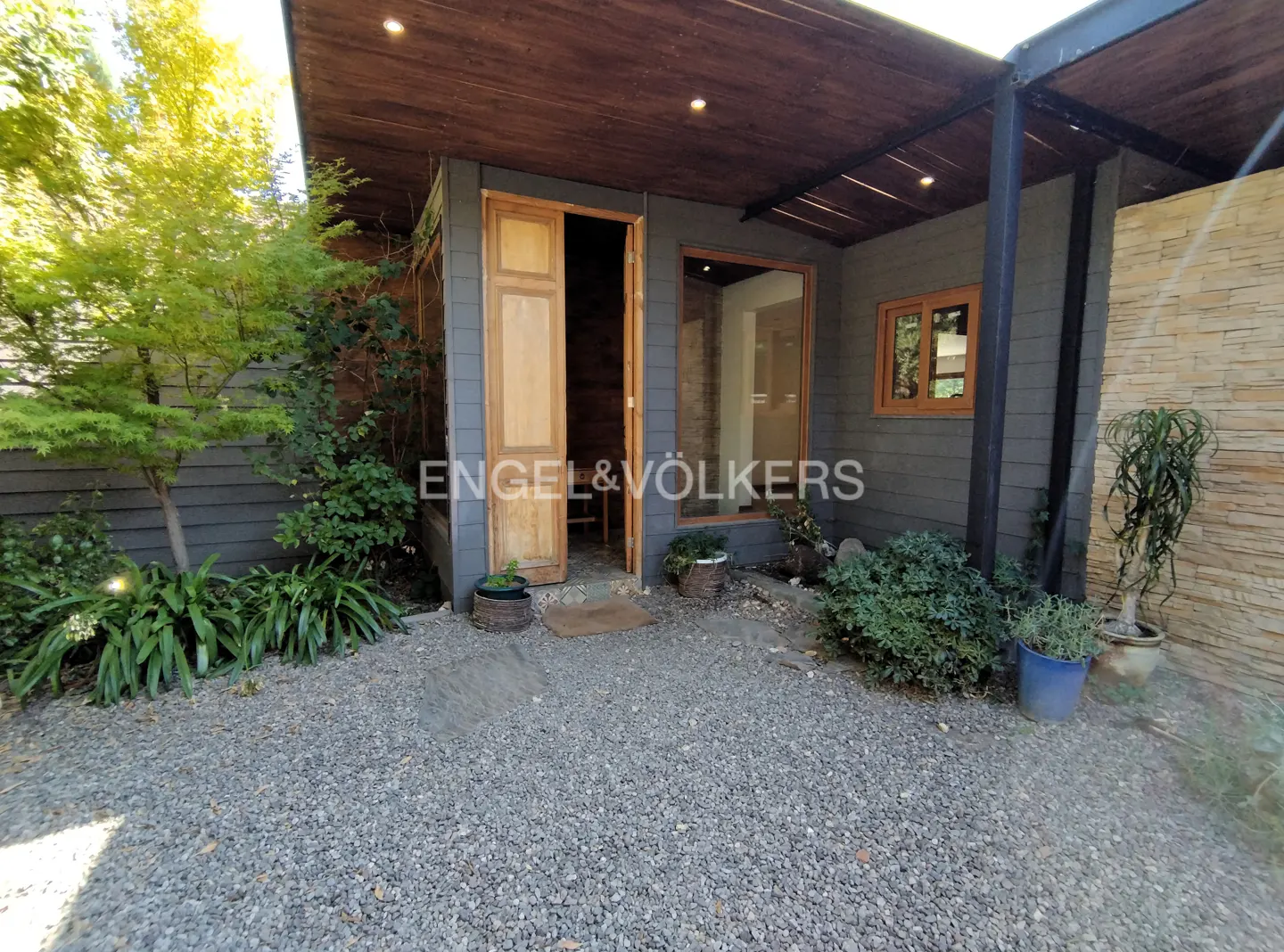 Exterior view of a gray house with an open wooden door, gravel yard, and green plants.
