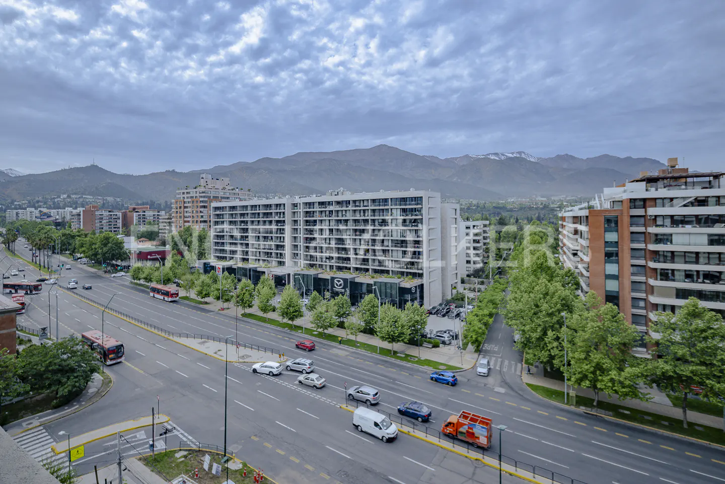 Cityscape view with apartment buildings, a busy street with cars and buses, and mountains in the background under a cloudy sky.