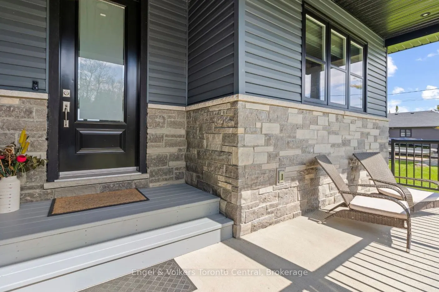 Exterior view of a house with gray siding, stone accents, and a black front door with a glass panel. Two lounge chairs sit on the porch.