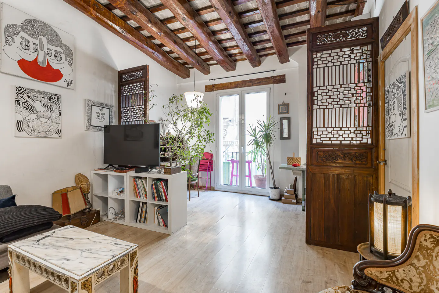 Living room with white walls, wooden beamed ceiling, and art. A TV sits on a white shelf. Balcony with pink stools visible through glass doors.