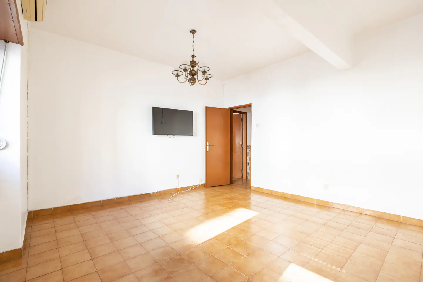 Bright, empty room with white walls, brown tile floor, TV, chandelier, and open brown door.