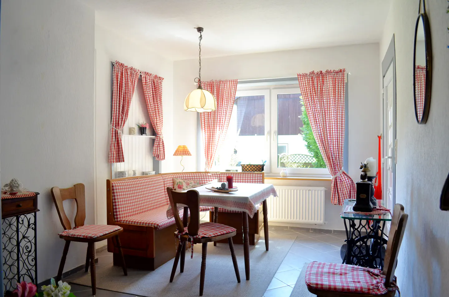 Cozy dining nook with a wooden corner bench, table, and chairs featuring red and white checkered cushions and curtains.