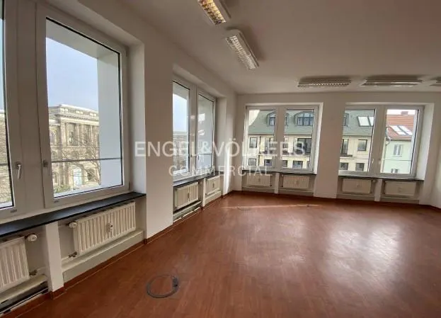 Empty office space with wood floors, white walls, and large windows overlooking city buildings. Radiators are under the windows.