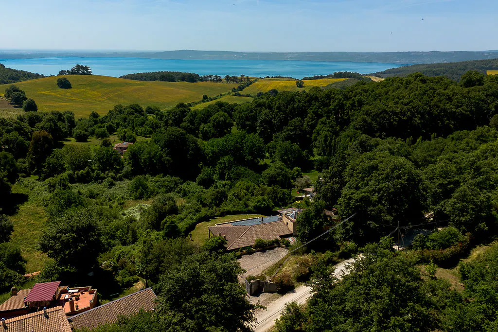 Aerial view of a rustic villa nestled in lush green hills overlooking a vast blue lake under a clear sky.