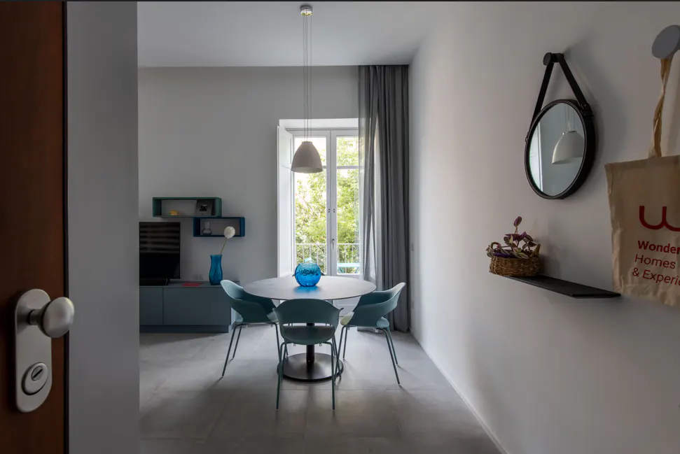 A bright, modern dining room with a round table, blue chairs, and a window view. A mirror and tote bag hang on the wall.
