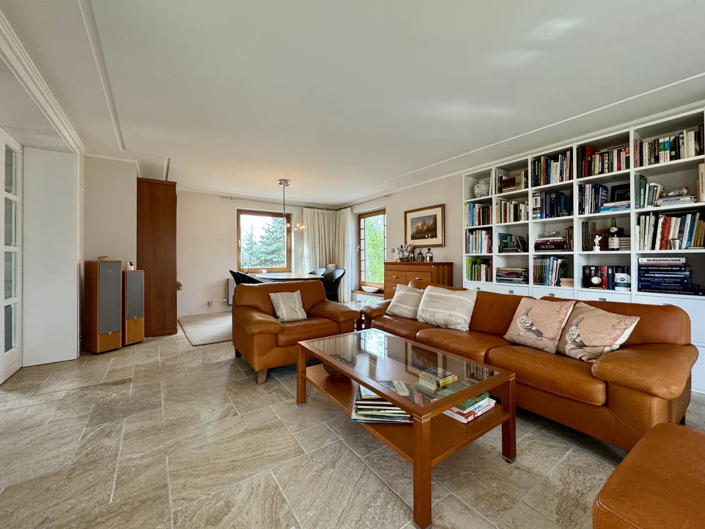 A living room with tan tile flooring, a brown leather sofa and armchair, and a wooden coffee table. A white bookcase filled with books is against the wall.