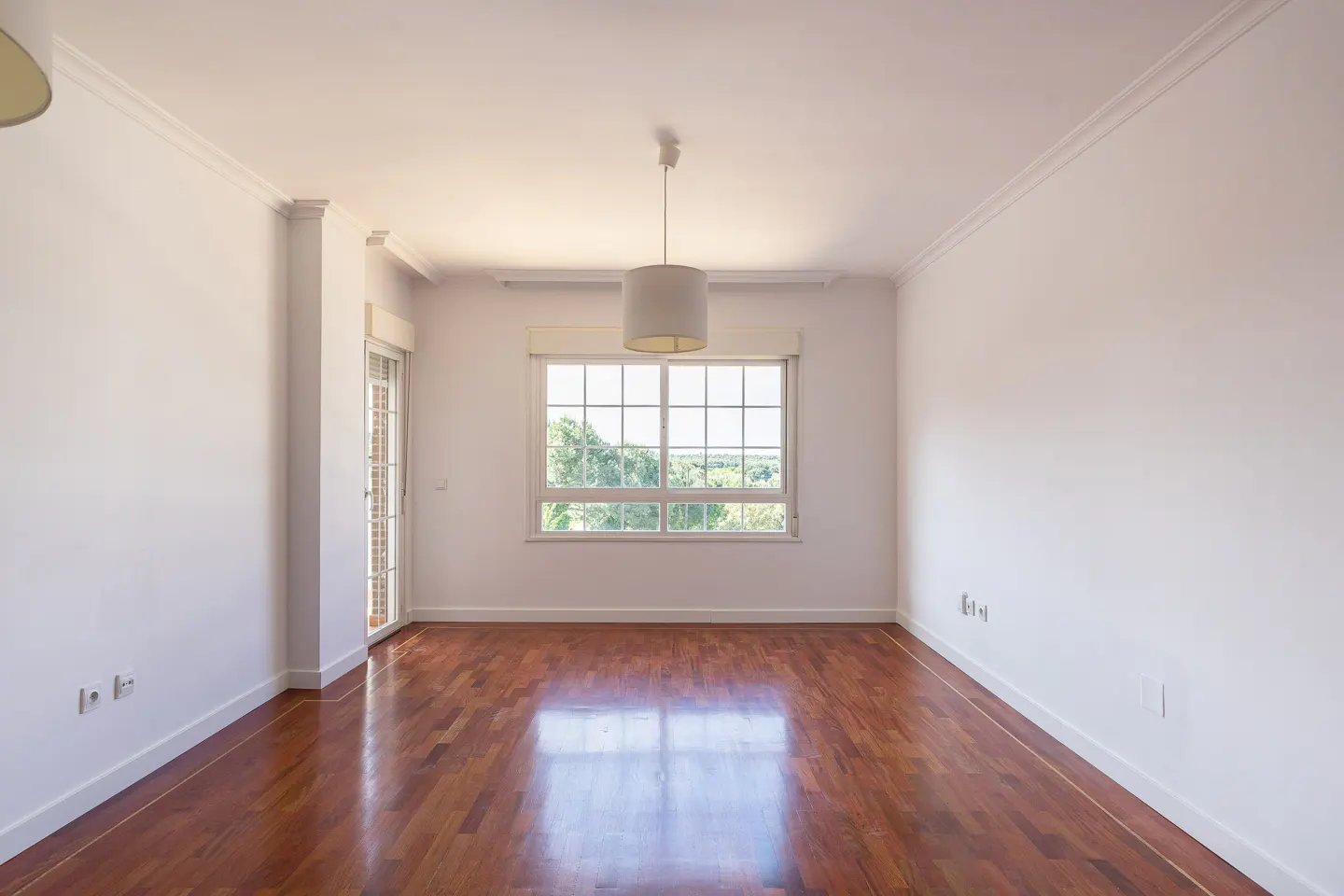 Empty room with white walls, wood floors, and a window with a view of trees. A white drum pendant light hangs from the ceiling.