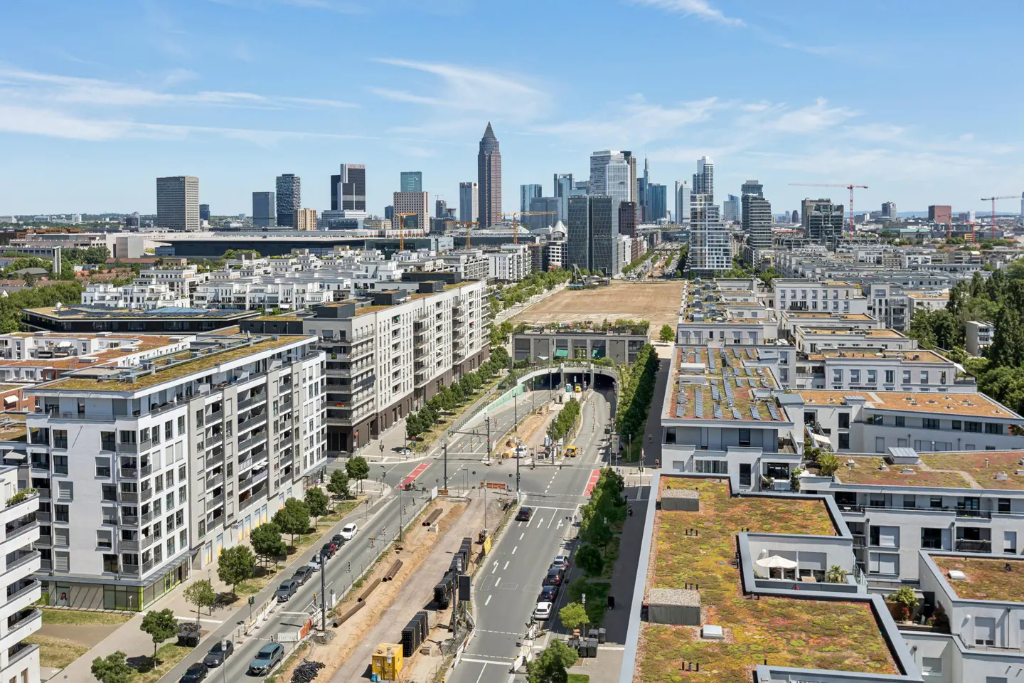 Cityscape view of Frankfurt, Germany, featuring modern white apartment buildings with green roofs, a road under construction, and skyscrapers in the background under a blue sky.