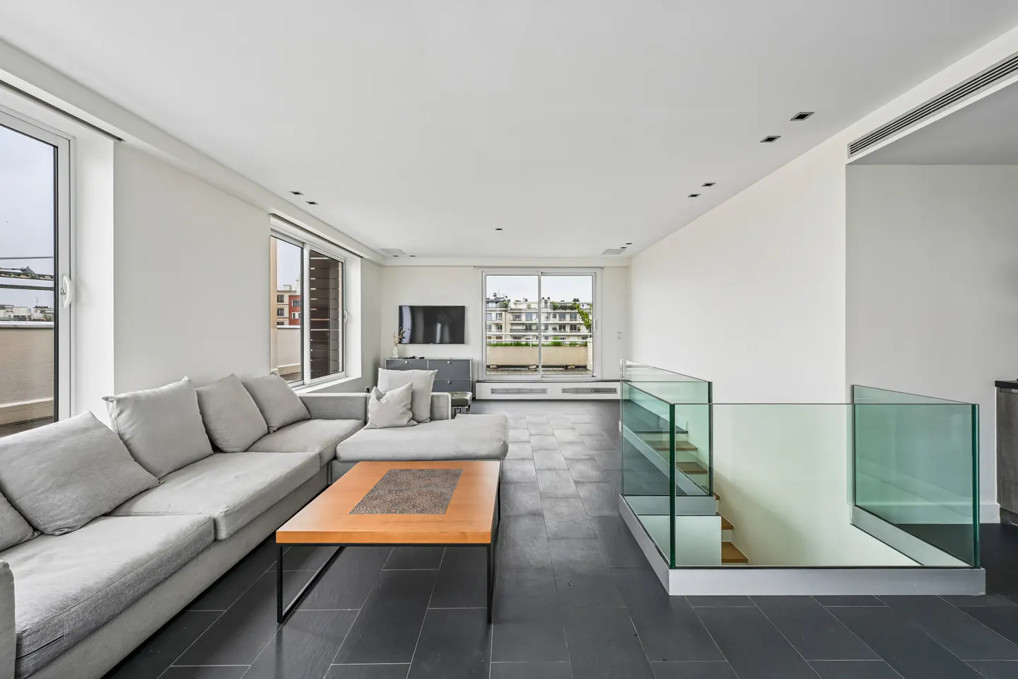 Bright living room with a gray sectional sofa, wood coffee table, and glass-railed staircase. Windows offer city views.