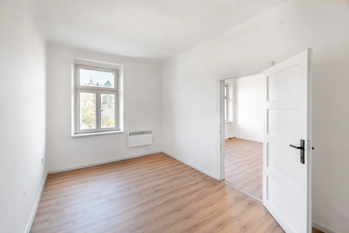 Bright, empty room with light wood floors and white walls. A window lets in natural light. An open white door leads to another room.
