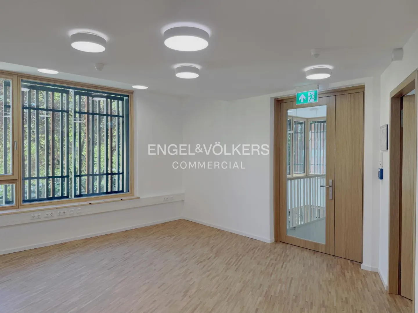 Bright, empty room with wood floors, white walls, and a large window with bars. Exit sign above a wooden door. Engel & Völkers logo in the center.