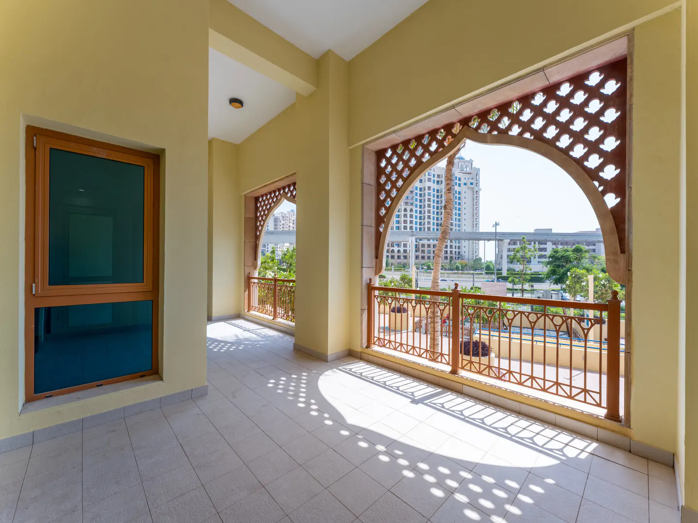 A bright, empty balcony with tan walls, tile floors, and decorative archways with views of buildings and greenery.
