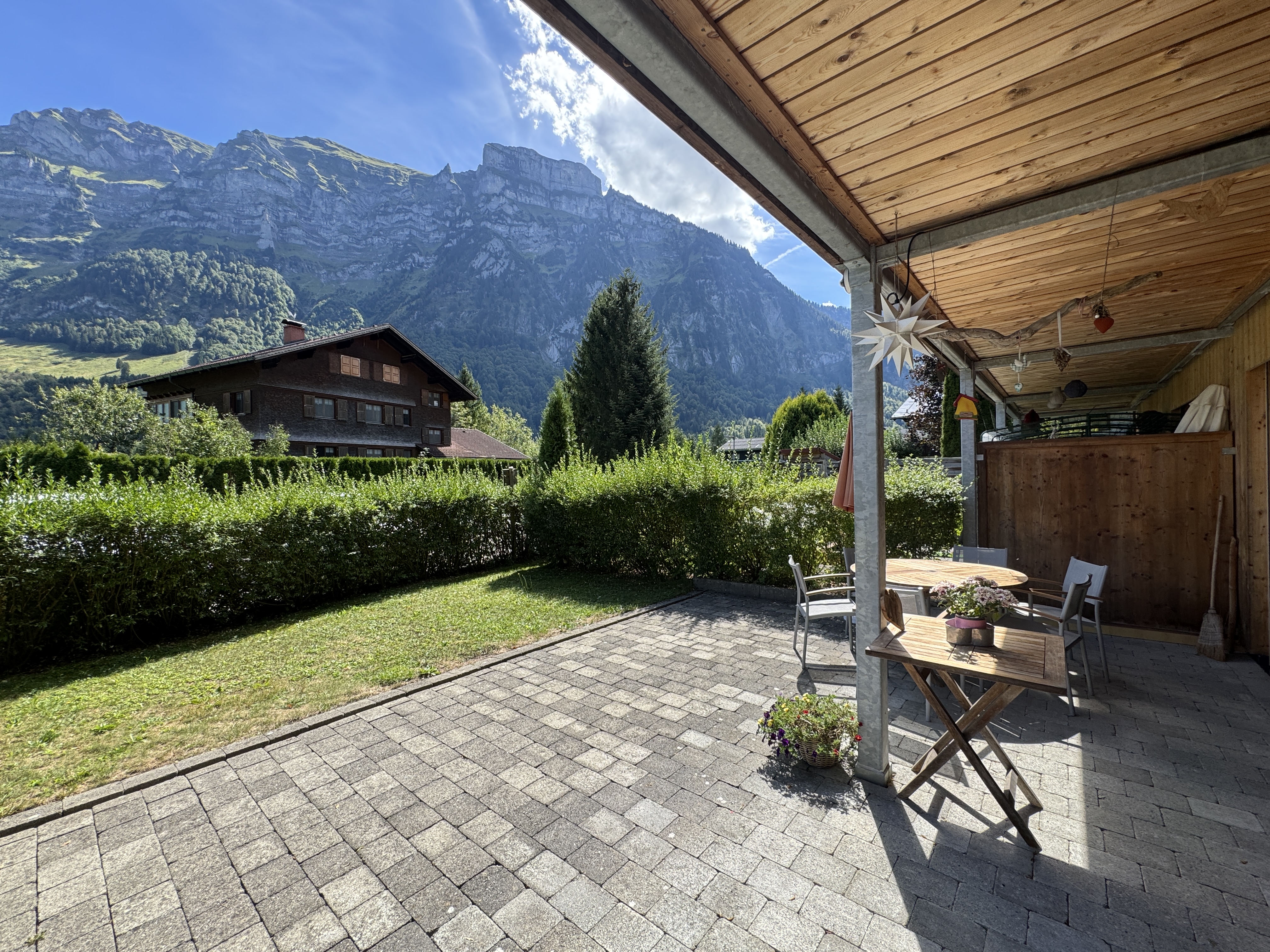 Covered stone patio with wooden ceiling, outdoor furniture, and a scenic view of a traditional house and mountain range. [AI-generated text]