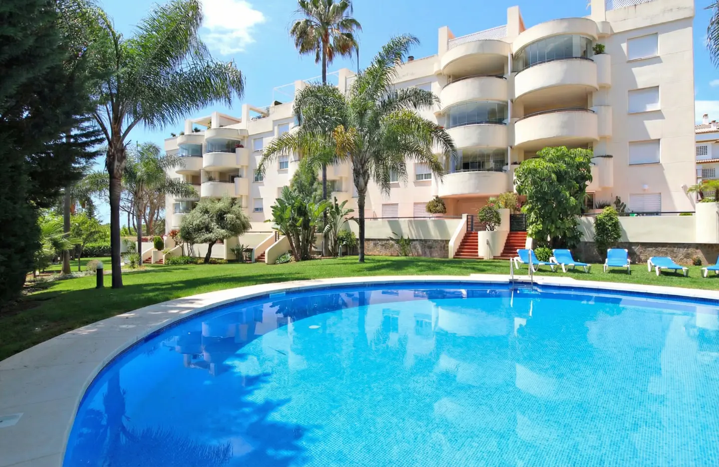 Exterior view of a multi-story apartment building with a blue swimming pool, green lawn, and palm trees on a sunny day.