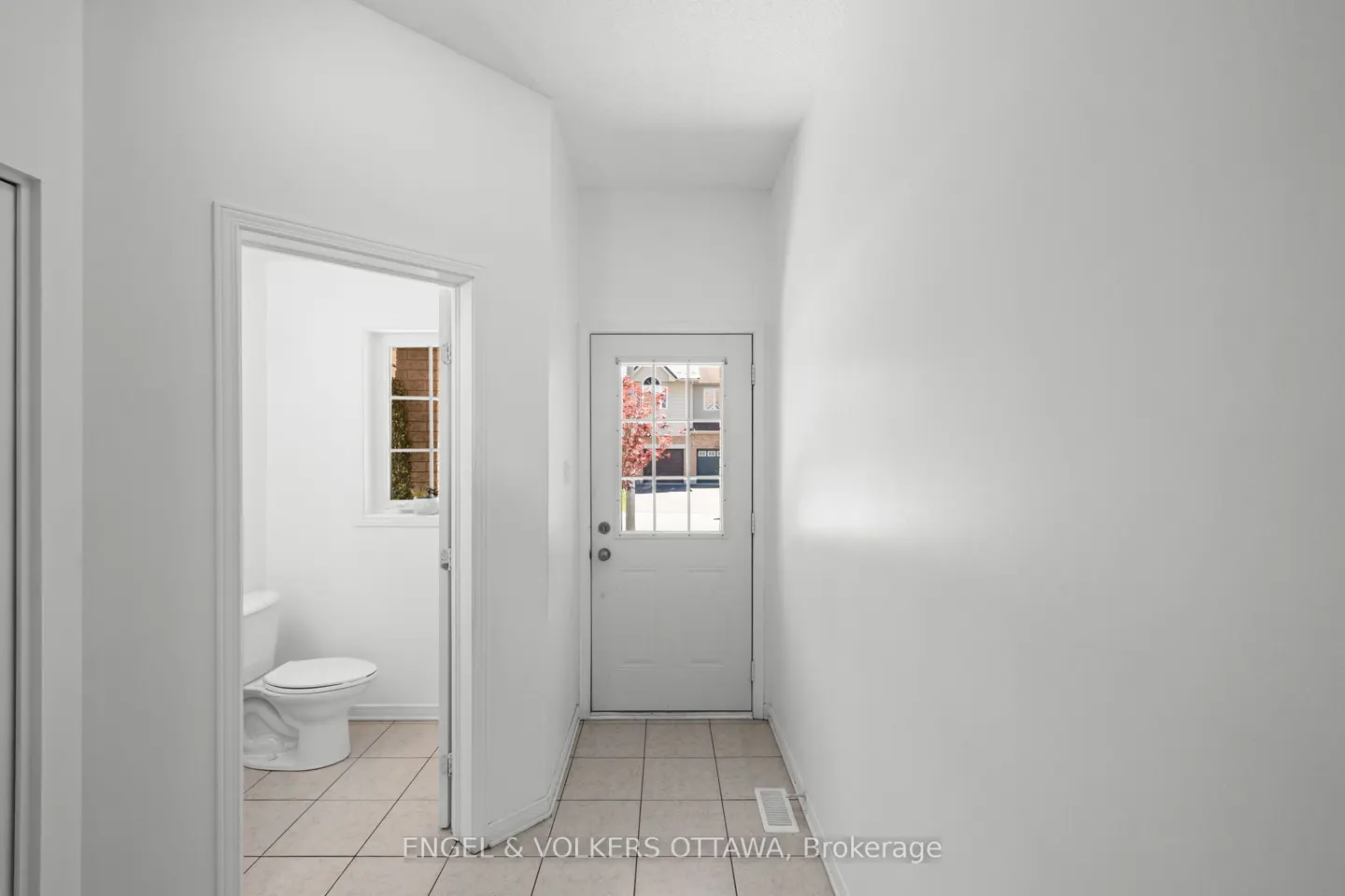 A bright hallway with white walls and tiled floors, featuring a white front door and an adjacent powder room.