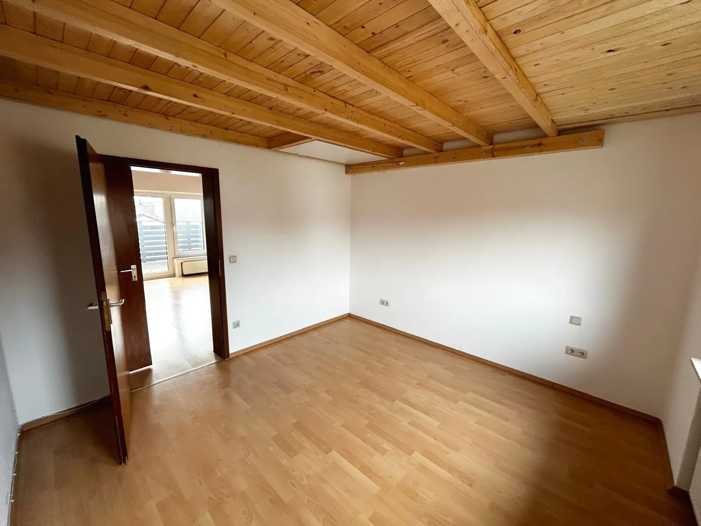 Empty room with light wood floors, white walls, and a wood plank ceiling. An open door reveals another room with a window.