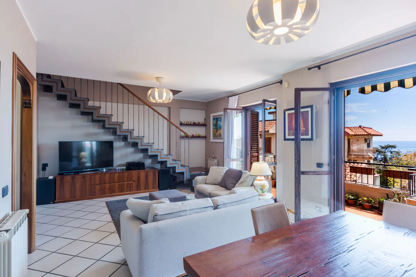 Living room with white sofas, wood table, TV, and stairs. Balcony doors open to a view of houses and the sea.