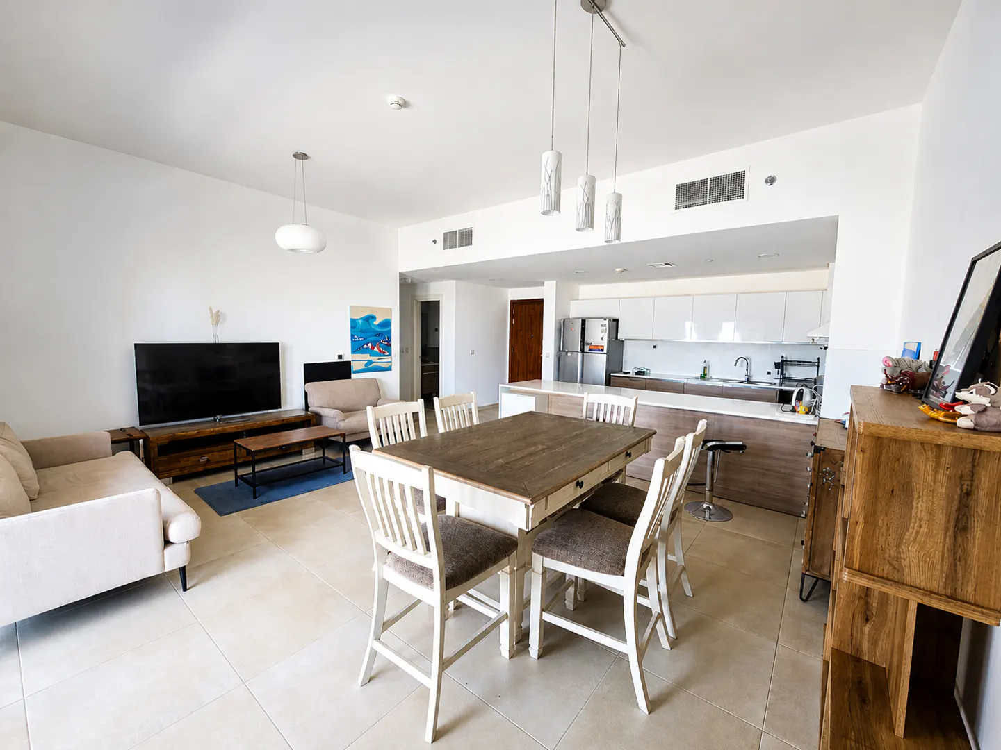 Bright, open-concept living space with a dining table, sofas, TV, and a modern kitchen with white cabinets.