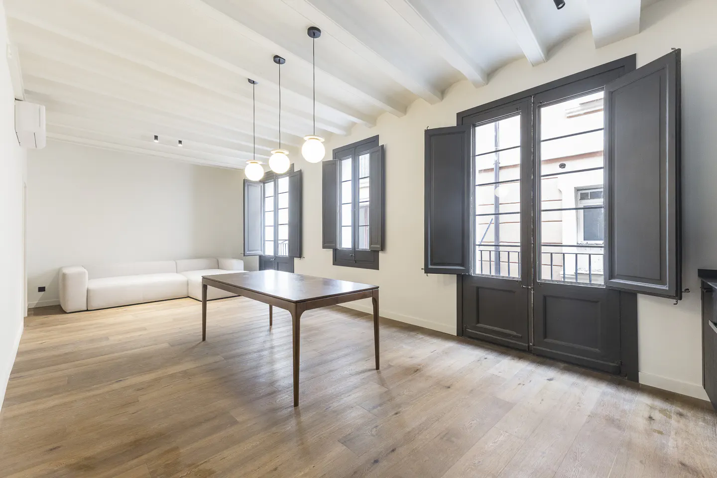 Bright living room with wood floors, white walls, and beamed ceiling. A wood table sits in the center, with a white sofa and dark windows.