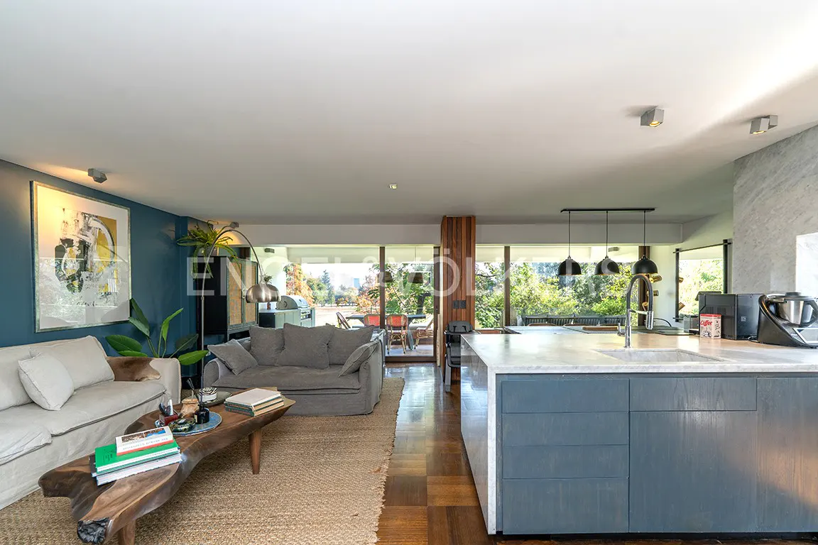 Open-concept living space with a blue kitchen island, sofas, and a wood coffee table on a jute rug. Large windows overlook a green outdoor area.