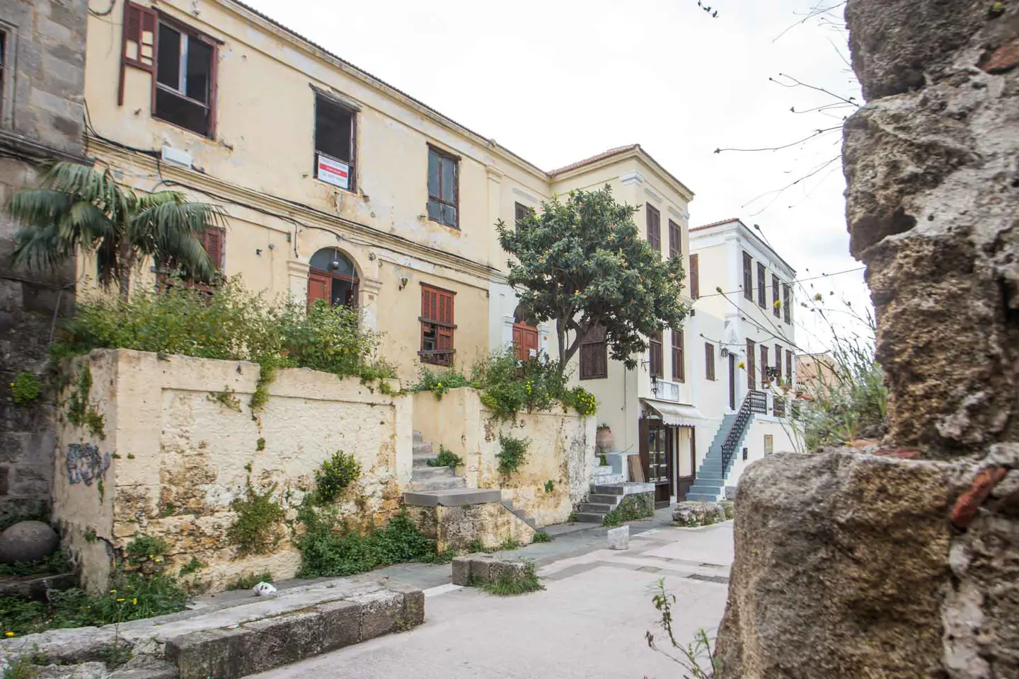 Exterior view of a yellow building with brown shutters and a "For Sale" sign, with stone walls and greenery.