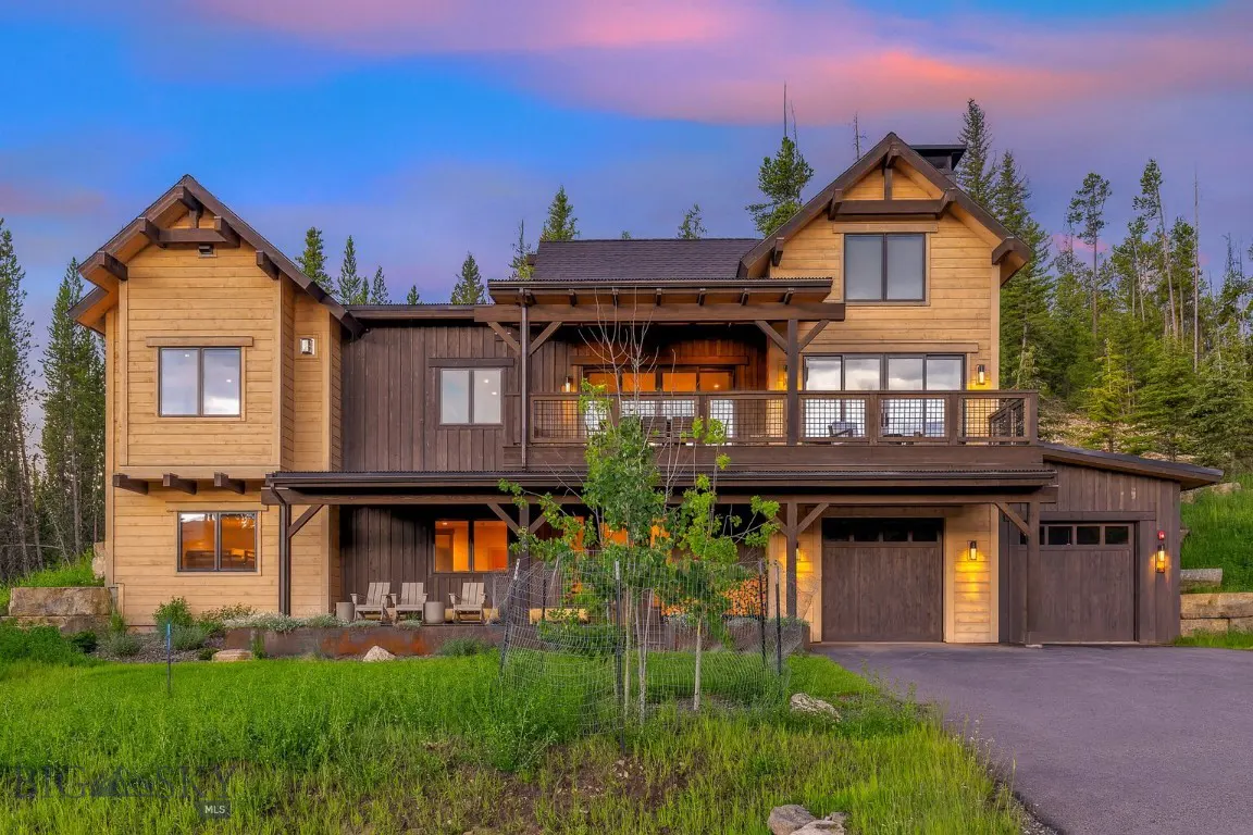 Two-story home with a balcony, brown wood siding, and a two-car garage. Green lawn and trees surround the house. Blue and pink sky.