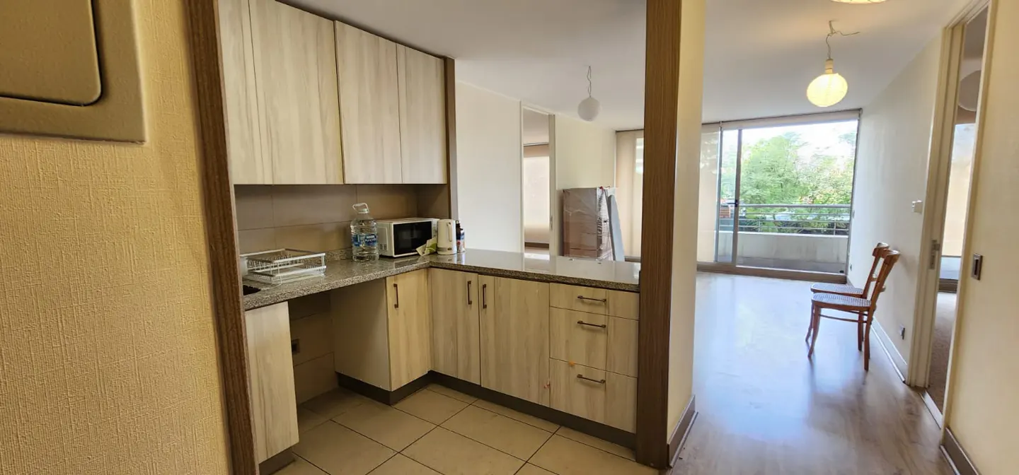 A bright apartment interior with light wood cabinets, a microwave, and a view of a balcony with trees.