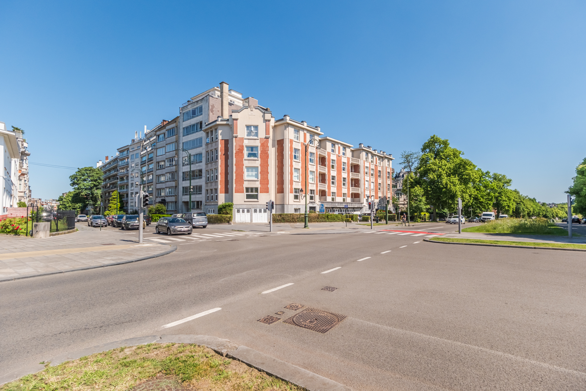 Street view of a multi-story apartment building with red brick accents and parked cars on a sunny day.