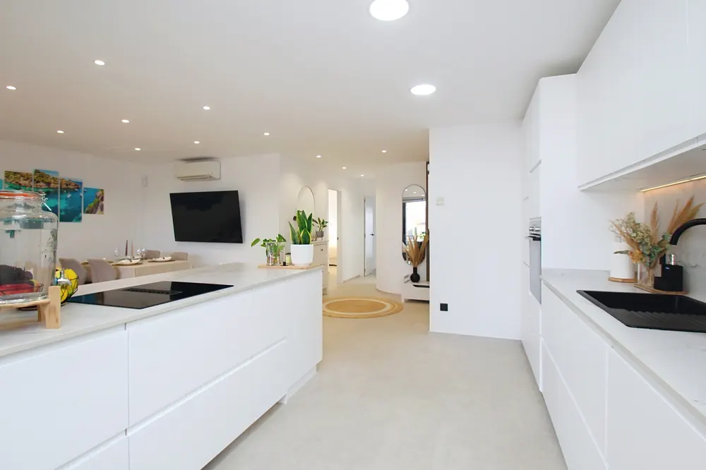 Bright, modern kitchen with white cabinets, countertops, and walls. A black sink and stovetop add contrast. Open floor plan leads to a dining area.