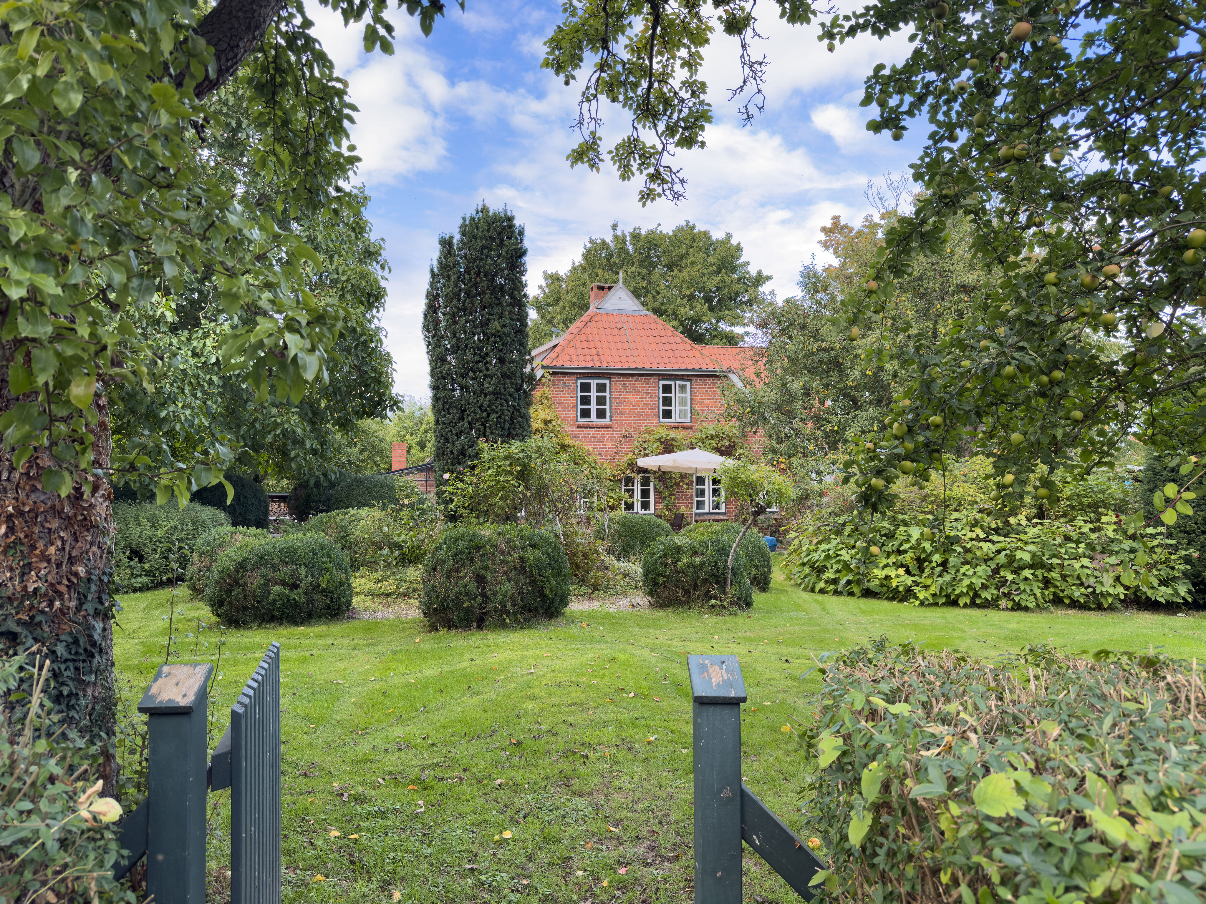 A red brick house with a tiled roof sits behind a lush green garden with manicured shrubs and trees under a cloudy sky. [AI-generated text]