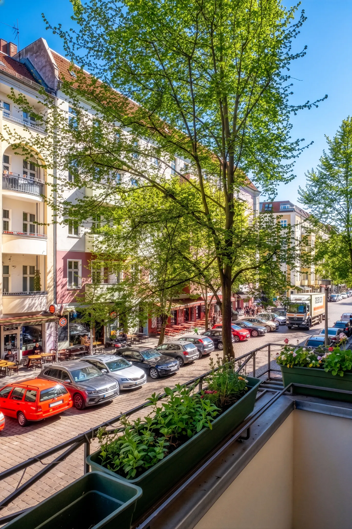View from a balcony with flower boxes overlooking a street with parked cars and buildings.