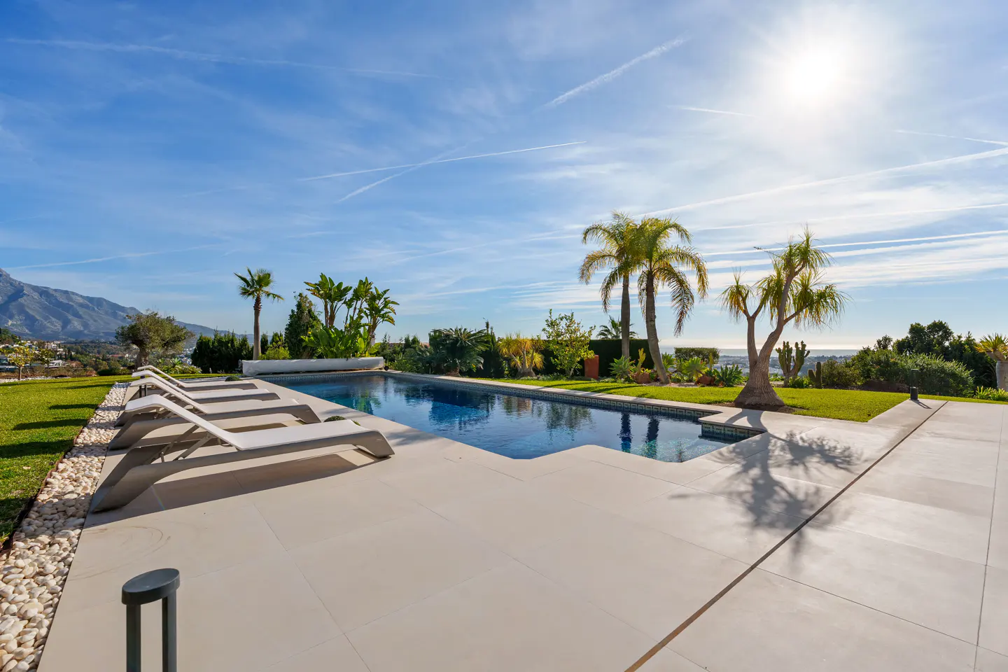 Outdoor pool with lounge chairs, palm trees, and mountain views under a blue sky.