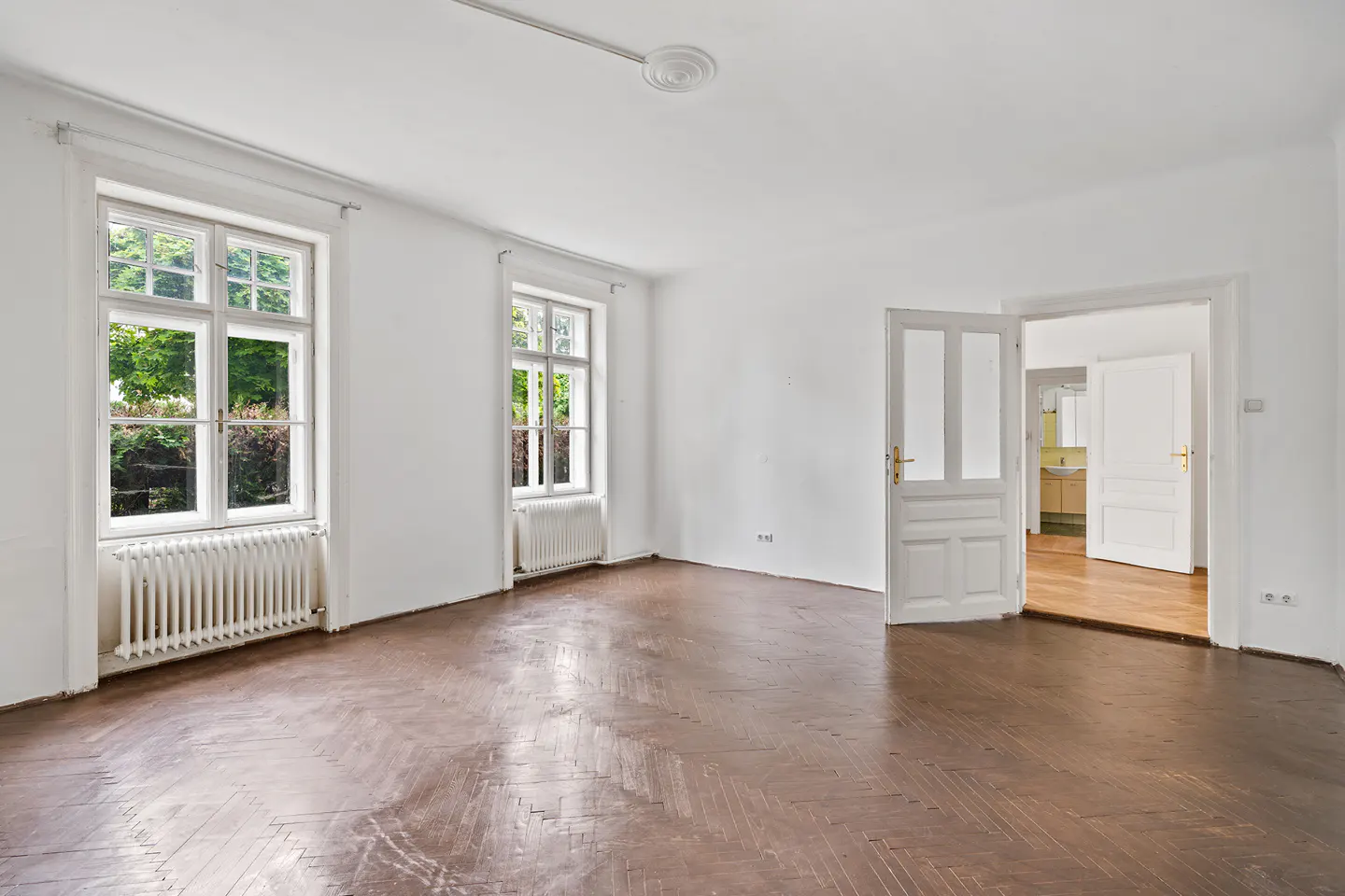 Bright, empty room with herringbone wood floors, white walls, two windows with radiators, and an open doorway to another room.