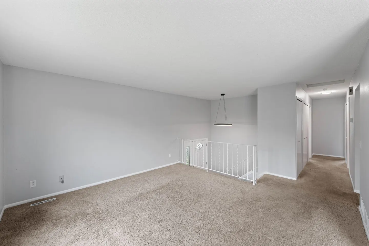 A bright, empty room with gray walls and beige carpet. A white railing separates the space from a hallway with closet doors. A modern light fixture hangs from the ceiling.