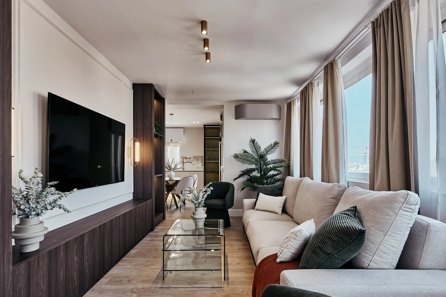 Living room with a large TV, beige sofa, glass table, and dining area in the background. Neutral tones and natural light.