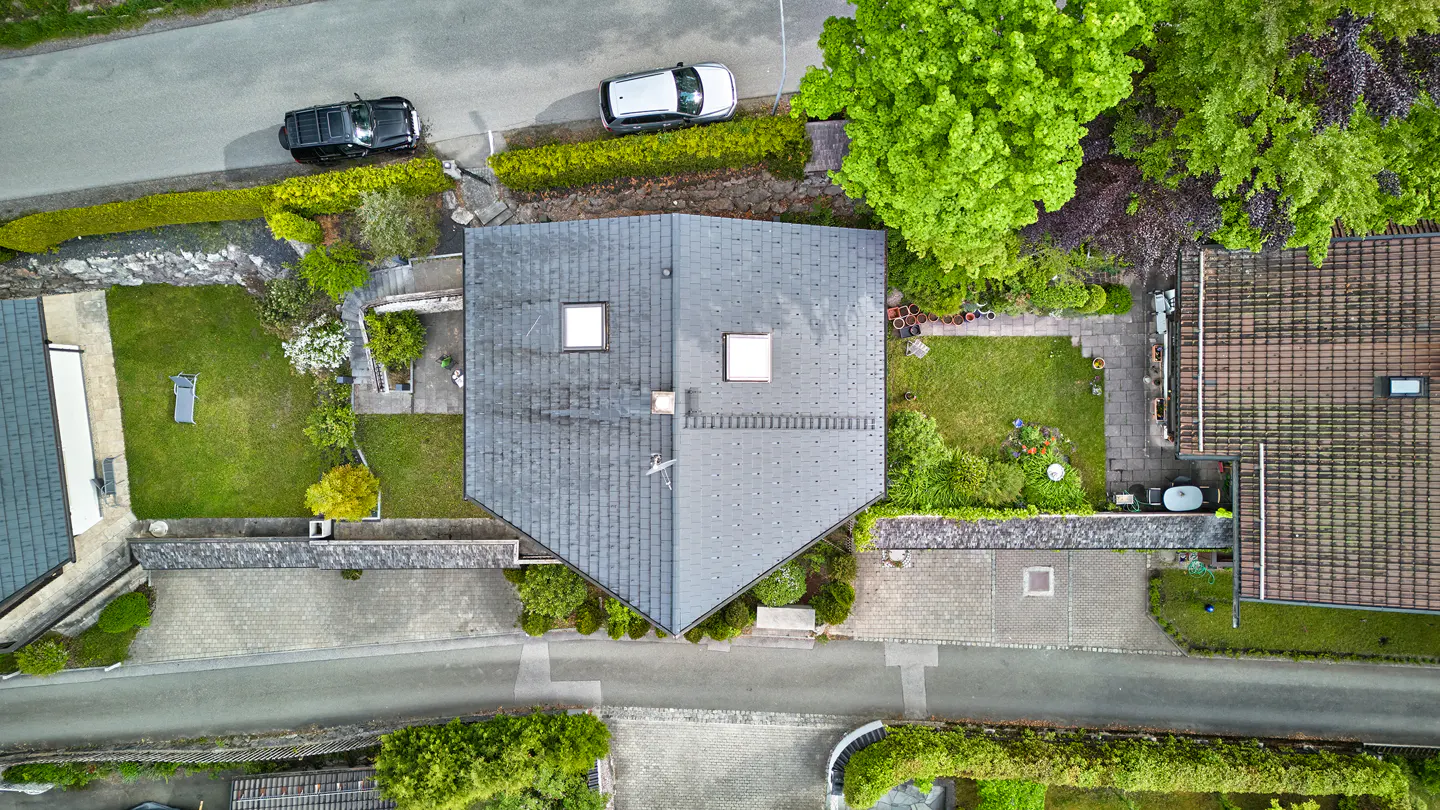 Aerial view of a house with a gray roof, skylights, green lawn, trees, and two parked cars.